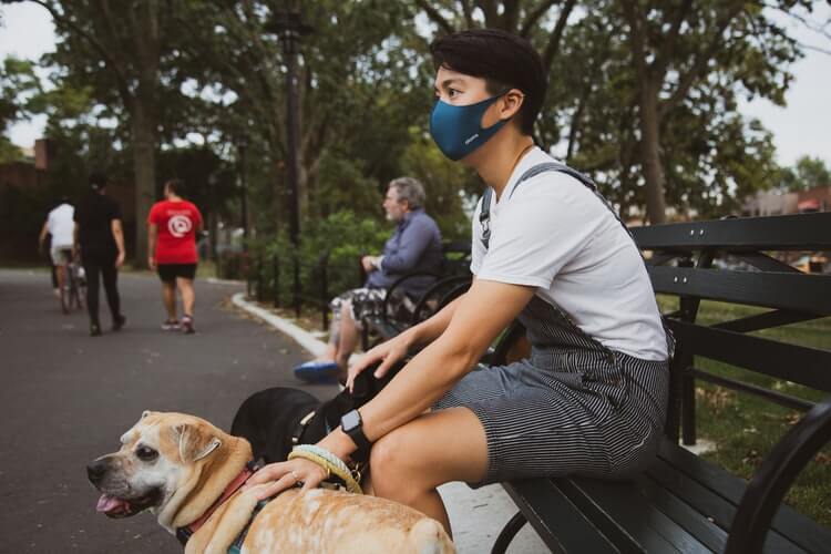 A man with a mask sitting on the bench in the park, his canine companion next to him