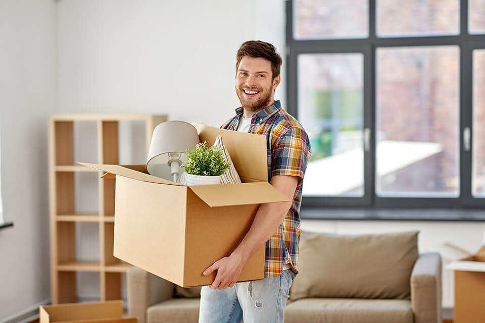 A smiling man holding a box with a lamp, plant, and a pillow