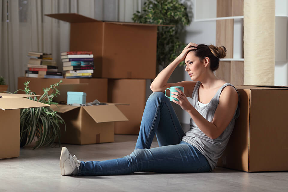 Worried woman sitting on the floor, holding her head and a mug, surrounded by boxes